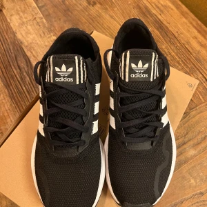 Svarta sneakers från Adidas - Snygga svarta sneakers från Adidas med klassiska vita ränder på sidan. Skorna har en bekväm vit sula och svart snörning. Perfekta för en stilren look. Bra för träning samt springa