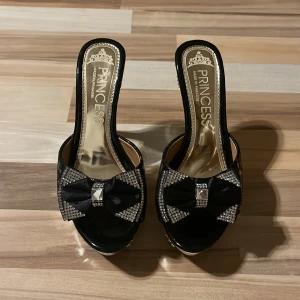 Svarta klackskor med rosett - Eleganta svarta klackskor från Princess med en glittrig rosett på ovansidan. Skorna har en genomskinlig klack och en öppen tå-design, vilket ger en stilren och festlig look.