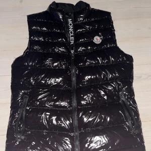 Svart dunväst från Moncler - Snygg svart dunväst från Moncler med glansig yta och dragkedja framtill. Västen har en hög krage och Moncler-logga på bröstet. Perfekt för kyligare dagar när du vill hålla stilen.