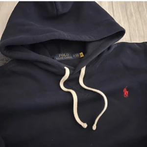Mörkblå hoodie från Ralph Lauren - Säljer en snygg mörkblå hoodie från Ralph Lauren med klassisk röd logga på bröstet. Den har en justerbar huva med vita snören och en bekväm passform. Perfekt för en avslappnad stil.