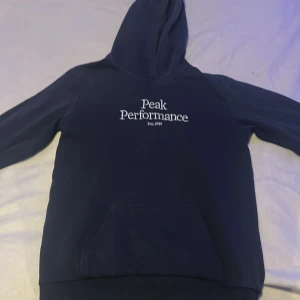Mörkblå hoodie från Peak Performance - Säljer en snygg mörkblå hoodie från Peak Performance med vit logga på bröstet. Tröjan har en klassisk känguruficka och en bekväm huva. Perfekt för kyliga dagar!