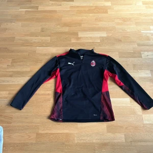 Svart och röd fotbollströja från Puma - Snygg svart och röd långärmad fotbollströja från Puma med AC Milan-logga på bröstet. Tröjan har en halv dragkedja och är perfekt för träning eller casual wear. Tillverkad i ett bekvämt material som andas.