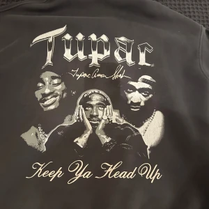  Grå svart hoodie med Tupac-tryck - Säljer en grå svart hoodie med Tupac-tryck och texten 'Keep Ya Head Up'. Hoodien har en stor ficka fram och justerbar huva med dragsko. Perfekt för dig som älskar klassisk hiphop-stil.