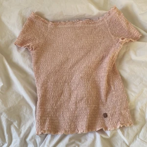 Rosa offshoulder topp - Säljer en söt rosa offshoulder topp med korta ärmar. Toppen har en smockad design som ger en stretchig och bekväm passform. Perfekt för varmare dagar!