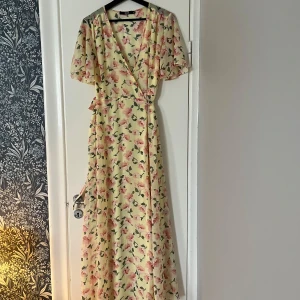 Blommig långklänning från Bik Bok - Säljer en vacker blommig långklänning i ljusgul färg med rosa blommor. Klänningen har en omlott-design med kortärmade volangärmar och ett knytband i midjan. Perfekt för sommarens alla tillfällen! 🌸