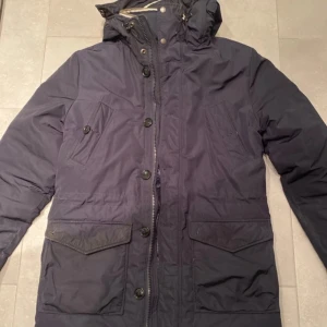 Mörkblå parkajacka från J.Lindeberg - Säljer en stilren mörkblå parkajacka från J.Lindeberg. Jackan har en avtagbar huva med pälsdetaljer och stängs med både dragkedja och knappar. Perfekt för kyliga dagar med sina praktiska fickor och långa ärmar.