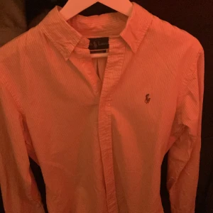 Randig skjorta från Ralph Lauren - Snygg randig skjorta från Ralph Lauren i en ljus orange nyans. Skjortan har långa ärmar och en klassisk krage med knappar framtill. Den är broderad med den ikoniska logotypen på bröstet. Perfekt för en stilren look.