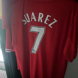 Röd Liverpool fotbollströja med Suarez 7 - Säljer en röd Liverpool fotbollströja från Warrior med Suarez och nummer 7 på ryggen. Tröjan har korta ärmar och klubbens emblem samt sponsorlogga i guld på framsidan. Perfekt för fans av Liverpool FC! I storlek M