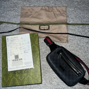 Svart axelväska från Gucci - Säljer en stilren svart axelväska från Gucci med ett diskret mönster och en praktisk dragkedja framtill. Väskan har en justerbar rem i rött och blått, vilket ger en snygg kontrast. Perfekt för att bära dina viktigaste saker med stil. Box samt kvitton finns