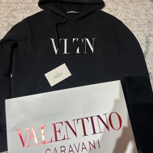 Svart hoodie från Valentino - Säljer en stilren svart hoodie från Valentino med VLTN-logga på bröstet. Tröjan har en klassisk design med huva och dragsko samt en praktisk magficka. Perfekt för en avslappnad och trendig look.