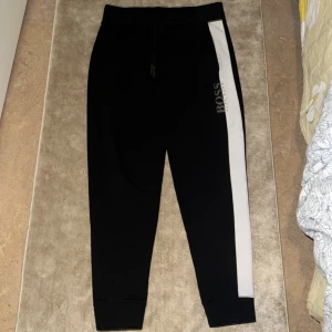 Svarta mjukisbyxor från Boss - Snygga svarta mjukisbyxor från Boss med en vit sidopanel och logotyp på benet. Byxorna har en elastisk midja med snörning för extra komfort. Perfekta för en avslappnad stil.