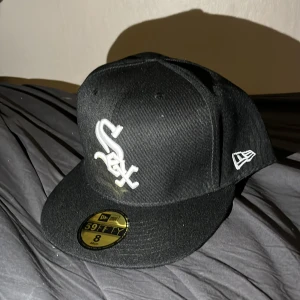 Svart keps från New Era - Snygg svart keps från New Era med broderad logga framtill. Kepsen är en 59FIFTY-modell med en platt skärm och en klassisk passform. Perfekt för att komplettera din streetwear-look. Helt oanvänd då den var för stor.
