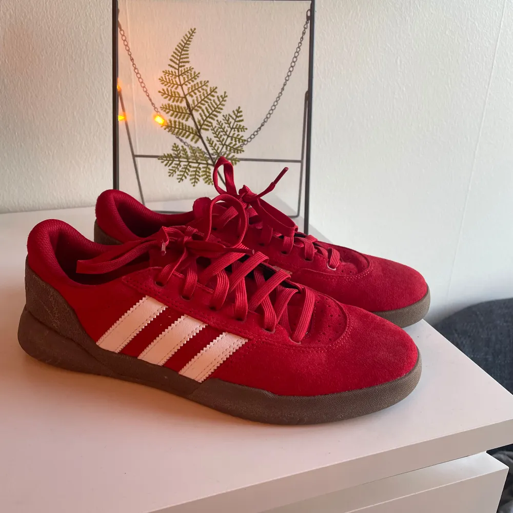 Säljer ett par snygga röda Adidas sneakers i mycket bra skick. De har den klassiska tre-randiga designen i vitt på sidan och är gjorda i mocka. Perfekta för både vardag och fest! Skorna har en bekväm passform och är i storlek herr 41 1/2. . Skor.