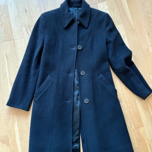 Svart kappa från Tiger of Sweden - Säljer en elegant svart ull kappa från Tiger of Sweden i mycket bra skick. Den har en klassisk design med långa ärmar och knappar framtill. Perfekt för höst och vinter, och ger en stilren look till vilken outfit som helst. Lätt figursydd. 