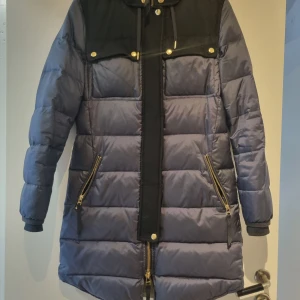 J. Lindeberg jacka  - Säljer en snygg dunjacka med huva. Jackan har långa ärmar och dragkedja framtill. Den är perfekt för kalla vinterdagar och har praktiska fickor med dragkedjor. Guldiga knappar ger en extra touch. Insidan är fodrad i grönt för extra värme. Luva är avtagbar. Jackan är använd en säsong och är i extremt bra skick. 