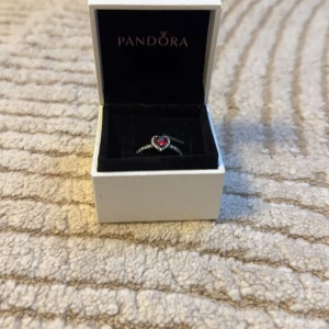 Ring med röd sten från Pandora - Säljer en elegant ring från Pandora med en vacker röd hjärtformad sten i mitten. Ringen är i silver och har en glittrig design som verkligen fångar ögat. Perfekt för speciella tillfällen eller som en fin present. Kommer i originalask.
