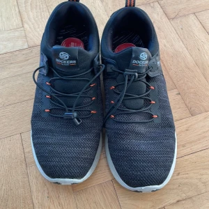 Svarta sneakers från Dockers - Säljer ett par mörkgrå  och svarta sneakers från Dockers i mycket bra skick. Tyget är grovt stickat. De har en ovandel med snörning och orange detaljer. Perfekta för både vardag och träning. Sköna och stilrena med en modern look. Använda ett fåtal gånger.