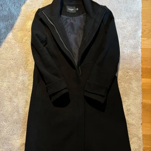 Svart kappa från Soaked in Luxury - Snygg svart kappa från Soaked in Luxury i storlek S. Perfekt för höst och vinter med sin stilrena design och långa ärmar. Kappan har en diskret dragkedja. Fodret i fickan på vänster sida är trasigt i sömmen men går lätt att laga om man har en symaskin. 