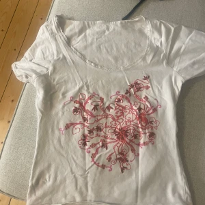 Vit t-shirt med rosa mönster - Säljer en söt vit t-shirt från AH Ling med ett rosa, snirkligt mönster framtill. T-shirten har korta ärmar och en rund halsringning med spetsdetaljer. Perfekt för en avslappnad sommarlook!