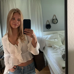 Vit blus med volangdetaljer - Säljer en superfin vit blus med volangdetaljer. Den är croppad och har långa ärmar, perfekt för både vardag och fest. Blusen är i ett lätt och luftigt material, vilket gör den idealisk för vår och sommar. Passar perfekt med jeans eller kjol för en chic look!