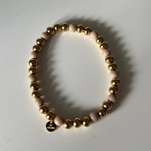 Guld och beige armband från By Jolima - Snyggt elastiskt armband med växelvis guld- och beige pärlor. Perfekt för att ge en elegant touch till vilken outfit som helst. Märkt med en liten rund bricka med 'By Jolima'.