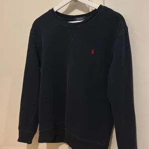 Blå Ralph Lauren tröja - Säljer en stilren blå tröja från Polo Ralph Lauren med det klassiska röda logotypbroderiet på bröstet. Tröjan har långa ärmar och är perfekt för både vardag och lite finare tillfällen. Superbekväm och i mycket bra skick!