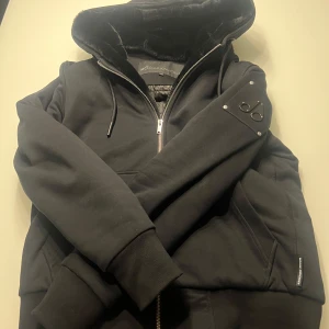 Moose Knuckles Hoodie/Jacka - Kvitto finns kan fraktas men står inte för frakten. Som ny knappt använd (Storlek M). Säljer för den inte passar mig längre köpte den när vintras tog slut förra året och sedan gick ner i vikt fick aldrig chans att använda. Kan frågas för fler bilder och bild på kvitto och hur många frågor som helst allt är äkta. Inga solskador den har varit upphängd i garderoben.