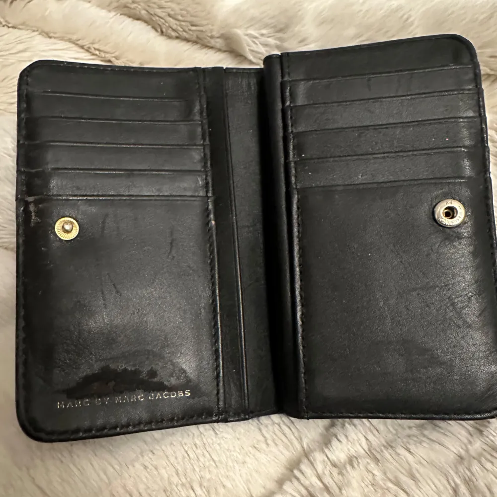 Säljer en elegant svart plånbok från Marc Jacobs i skinn. Den har flera kortfack och en snygg guldfärgad logotyp på framsidan. Perfekt för att hålla ordning på kort och kontanter med stil. Plånboken är i bra skick, dock lite skavd på insidan - se bild 2, och har en klassisk design som passar alla tillfällen. Inköpt 2019. . Asusteet.