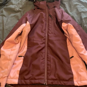 Skidjacka - Säljer en snygg lila och rosa jacka från Ski Industries i storlek XS. Den är vind- och vattenavvisande, perfekt som tredje lager för skidresor. Jackan har dragkedja och en praktisk huva. Perfekt för vinterns äventyr! ❄️ endast använd 4 dagar i sälen