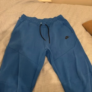 Blåa Nike tech byxor  - Säljer ett par blå mjukisbyxor från Nike i storlek L. Byxorna har en bekväm passform med snörning i midjan och är perfekta för avslappnade dagar. De är i acceptabelt skick med en liten reva på ena benet. Perfekta för chill eller träning