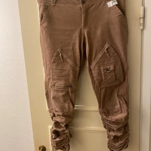 Culture cargopants med dragkedjor - Säljer ett par bruna cargopants i bra skick. De har flera praktiska fickor med dragkedjor och knappar, perfekt för en casual look. Byxorna är loose fit och passar bra för höst och vår. Materialet känns slitstarkt och bekvämt. Benen veckade nedtill med gummiband.