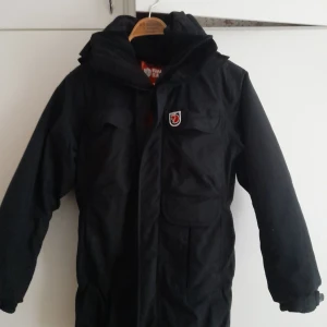 Svart parkajacka från Fjällräven - Säljer en svart parkajacka från Fjällräven i storlek XXS. Jackan har en praktisk huva och flera fickor framtill. Perfekt för kalla vinterdagar med sin robusta design och varma material. Den är i mycket bra skick och redo för nya äventyr!