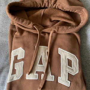 Brun hoodie från GAP - Säljer en brun hoodie från GAP i mycket bra skick. Den är supermjuk och varm med fleecefoder, perfekt för höst och vinter. Har en stor ficka fram och justerbar huva. GAP-loggan är i beige på bröstet. Perfekt för mysiga dagar eller casual outfits! Finns en liten svag fläck på fickan, se bild. Syns knappt.