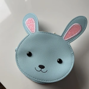 Söt kaninformad axelväska för barn  - En supergullig axelväska i form av ett kaninansikte. Väskan är ljusblå med rosa detaljer i öronen och har svarta ögon och nos. Perfekt för att lägga småsaker i och ha med sig på äventyr. Den är gjord av syntetmaterial och har en dragkedja för säker förvaring.