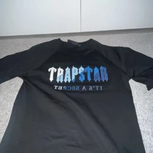 Svart t-shirt från Trapstar - Säljer en svart t-shirt från Trapstar med coolt tryck i blått och vitt på framsidan. Perfekt för en avslappnad stil och passar till alla tillfällen. T-shirten är i bra skick och har en normal passform. Skön och stilren, ett måste i garderoben!