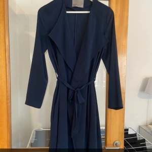 Mörkblå trenchcoat - Snygg mörkblå trenchcoat med knytbälte i midjan. Perfekt för höst och vår. Jackan har långa ärmar och en elegant, flytande design. Passar både till vardags och festliga tillfällen.