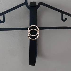 Svart bälte med gulddetaljer - Snyggt svart bälte med dubbla guldringar som spänne. Perfekt för att ge en stilren touch till vilken outfit som helst. Tillverkat i Kina och mäter 65 cm i längd. Passar både till vardags och festliga tillfällen. 🖤✨