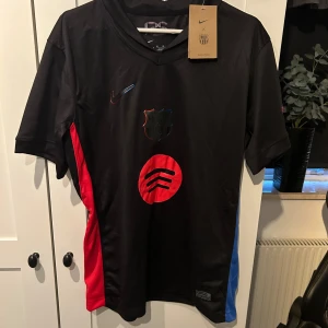 FC Barcelona fotbollströja från Nike - Säljer en svart FC Barcelona fotbollströja från Nike i nytt skick. Den har korta ärmar och coola detaljer i rött och blått på sidorna. Märken och loggor på framsidan ger den en unik look. Perfekt för både träning och vardag!