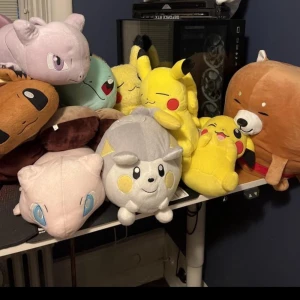 Samling av Pokémon-gosedjur - En samling av stora och mjuka Pokémon-gosedjur, inklusive populära karaktärer som Pikachu och Mew. Perfekt för Pokémon-fans som vill ha en mysig och färgglad dekoration i rummet. Gosedjuren är i olika färger och storlekar, vilket gör dem till en rolig och varierad samling.