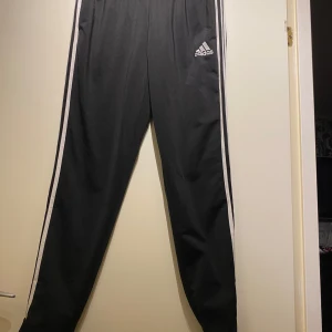 Svarta träningsbyxor från Adidas - Snygga svarta träningsbyxor från Adidas med klassiska vita ränder längs sidorna och logotyp på låret. Byxorna har en elastisk midja för extra komfort. Perfekta för sport och fritid. Omkrets midja: 81 cm Beninnerlängd: 78 cm Längd utsida ben: 106 Smala ben Resår i midja och muddar i beslut  Inga bilder när den sitter på mig! Katter finns i hemmet