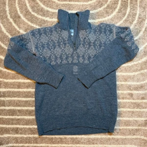 Blå stickad tröja från Snjor - Säljer en snygg blå stickad tröja från Snjor med ett klassiskt mönster i ljusare nyanser. Tröjan har en praktisk dragkedja vid halsen och långa ärmar. Perfekt för kyliga dagar!