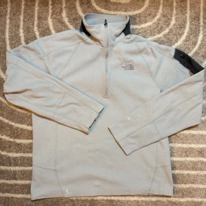 Grå fleece tröja från The North Face - Säljer en grå fleece tröja från The North Face med halv dragkedja och svarta detaljer på axlarna. Perfekt för kyliga dagar och outdoor-äventyr. Tröjan har långa ärmar och en bekväm passform. Har små defects men påverkar inte looken 