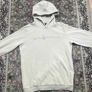 Sail racing hoodie - Säljer en stilren vit hoodie från Sail Racing med tryck på bröstet. Den har en klassisk känguruficka och en bekväm huva. Perfekt för en avslappnad stil. Använd 1-2 gånger Passar Xs,S