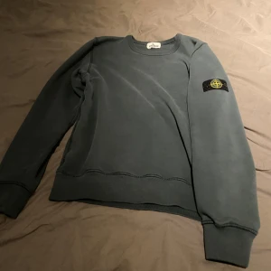 Grå sweatshirt från Stone Island - Säljer en stilren grå sweatshirt från Stone Island med deras ikoniska märke på ärmen. Tröjan har en klassisk rund hals och långa ärmar, perfekt för en avslappnad look. Passar bra till både jeans och joggers.
