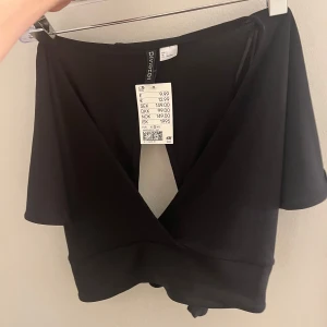 Svart omlottopp från H&M - Snygg svart omlottopp från H&M med knytning i ryggen. Toppen har en djup v-ringning och korta ärmar, perfekt för en stilren look.