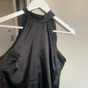 Svart halterneck topp - Elegant svart halterneck topp  i satin med hög hals och knytning i ryggen. Perfekt för en stilren look.