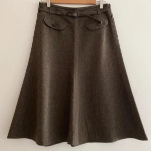 Tweed Brun kjol med bälte - Elegant brun kjol med bälte och dekorativa fickor framtill. Kjolen har en klassisk A-linjeform som ger en tidlös look. Perfekt för en stilren och sofistikerad outfit.