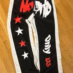 Svarta flared byxor med stjärnor och text - Coola svarta byxor med vita och röda detaljer. Byxorna har vita sidor och är dekorerade med röda och vita stjärnor samt text. Perfekta för en avslappnad stil.