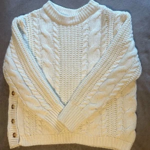 Vit stickad tröja med knappar - Mysig vit stickad tröja med flätmönster och dekorativa knappar på sidan. Perfekt för kyliga dagar med sin varma och bekväma design. Tröjan har långa ärmar och en rund halsringning.