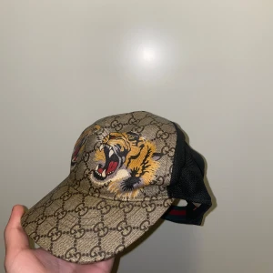 Gucci keps med tigerbroderi - Snygg keps från Gucci med ett djärvt tigerbroderi på framsidan. Kepsen har ett klassiskt Gucci-mönster i beige och brunt, med svarta meshdetaljer på baksidan för ventilation. Perfekt för att ge din outfit en lyxig touch.